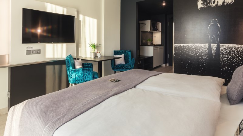 Modernes Zimmer mit großem Bett, blauen Sesseln, Astronauten-Wandbild, Flachbildfernseher und kleiner Küche., © Novum Management GmbH