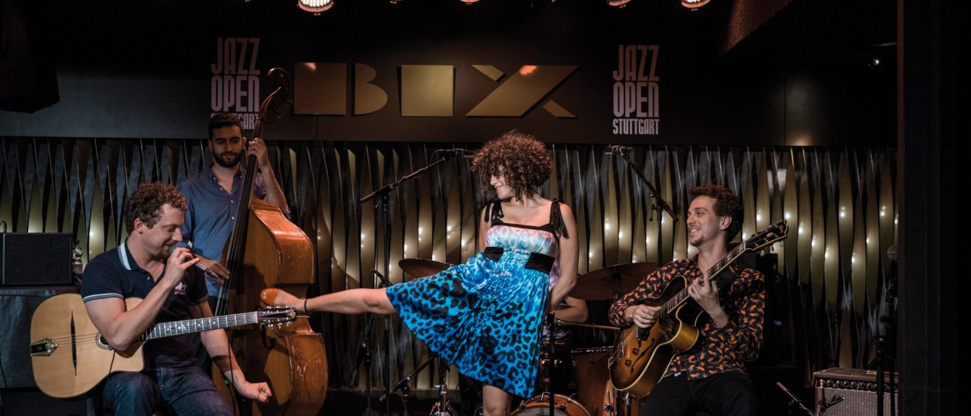 Eine Band spielt im BIX Jazzclub. Ein Gitarrist, ein Kontrabassist und eine Sängerin in blauem Kleid sind zu sehen. "Jazz Open Stuttgart" im Hintergrund., © Peter Steinheisser