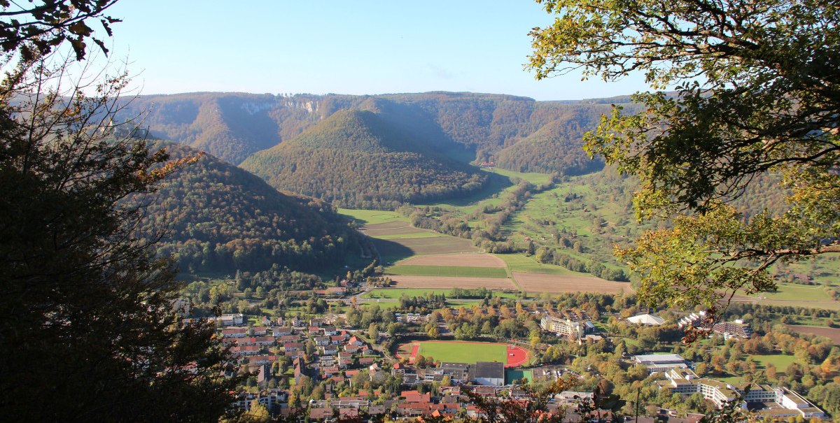 Ausblick vom Nägelesfelsen auf eine Stadt mit Sportplatz, umgeben von grünen Hügeln und Wäldern unter klarem Himmel., © Bad Urach Tourismus