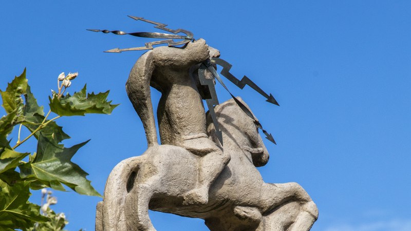 Steinfigur eines Reiters mit Blitzsymbolen, umgeben von grünen Blättern, vor klarem, blauem Himmel., © SMG, Achim Mende
