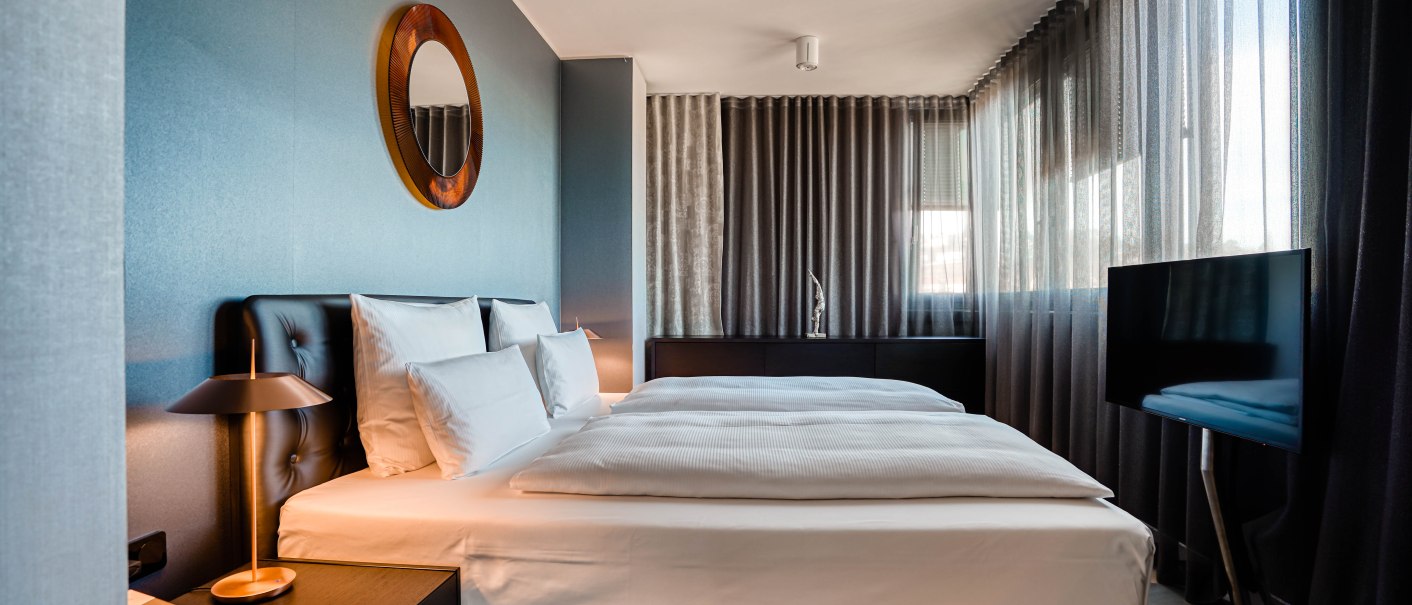 Modernes Hotelzimmer mit Doppelbett, Spiegel an der Wand, Nachttischlampen und Fernseher. Große Fenster mit Vorhängen., © CLOUD N°7 APARTMENTS GmbH
