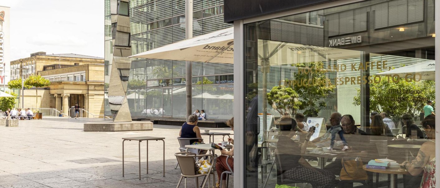 Menschen sitzen in einem Caf&eacute; auf einem st&auml;dtischen Platz. Moderne Geb&auml;ude und ein Kunstwerk sind im Hintergrund zu sehen., &copy; SMG, Sarah Schmid