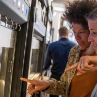 Zwei Frauen stehen vor einem Weinspender und zeigen auf verschiedene Weinflaschen. Im Hintergrund sind weitere Flaschen und Gl&auml;ser zu sehen., &copy; Stuttgart Marketing GmbH, Fotografin Martina Denker