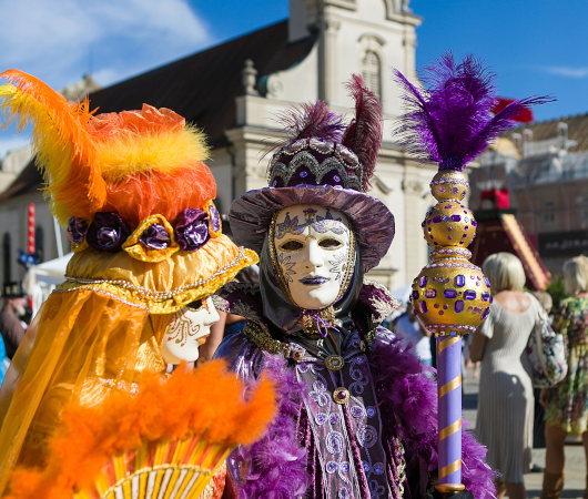 Zwei Personen in prächtigen venezianischen Kostümen und Masken stehen vor einer Kirche. Sie tragen bunte Federn und halten dekorative Accessoires., © Tourismus & Events Ludwigsburg Zwei Personen in prächtigen venezianischen Kostümen und Masken stehen vor einer Kirche. Sie tragen bunte Federn und halten dekorative Accessoires., © Tourismus & Events Ludwigsburg