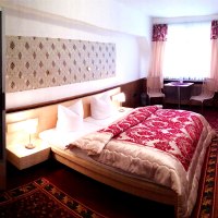 Gemütliches Hotelzimmer mit Doppelbett, rotem Teppich, Vorhängen, kleinem Tisch, zwei Stühlen und Fernseher.