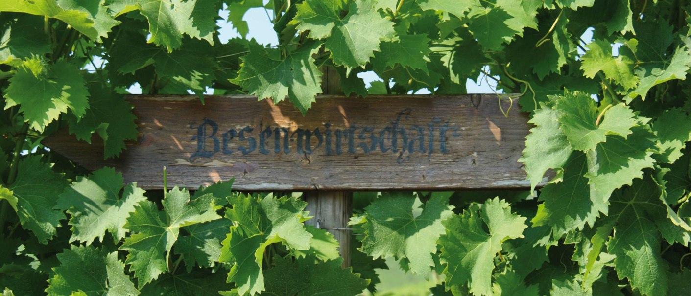 Ein Holzschild mit der Aufschrift 'Besenwirtschaft' ist von grünen Weinblättern umgeben., © SMG Ein Holzschild mit der Aufschrift 'Besenwirtschaft' ist von grünen Weinblättern umgeben., © SMG