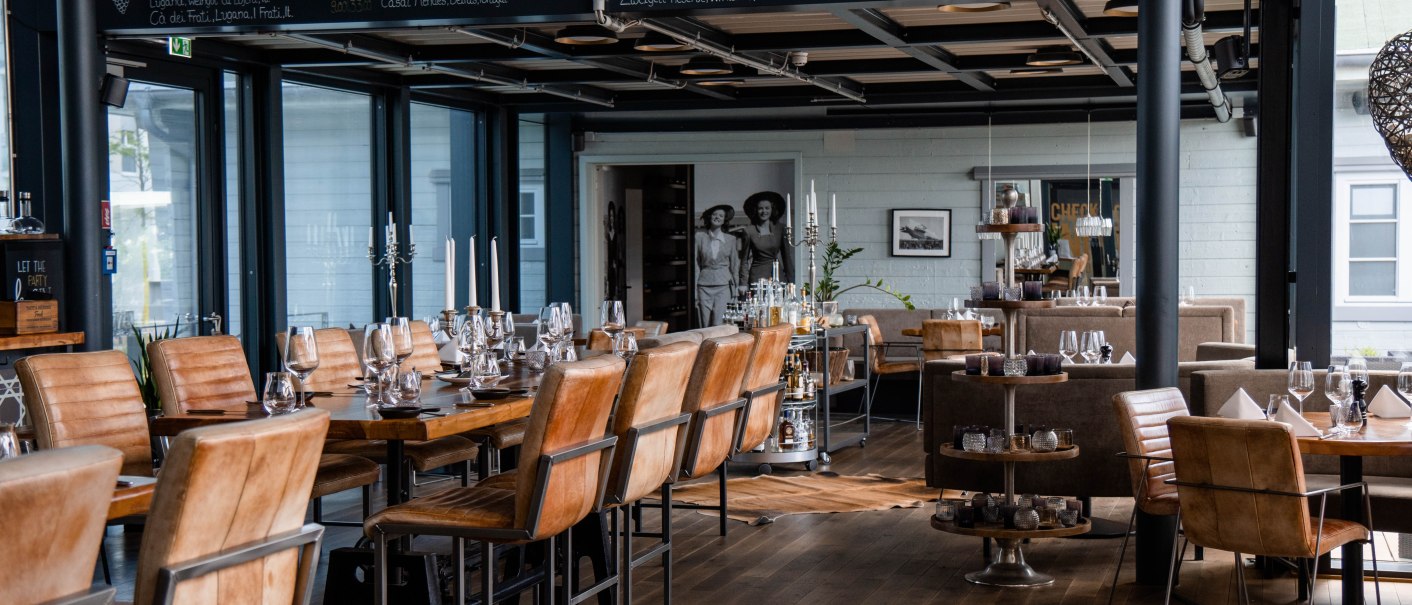 Stilvolles Restaurant mit Holzböden, Ledersesseln und gedeckten Tischen. An der Wand eine Tafel mit Weinauswahl. Elegante Atmosphäre., © Check Inn Restaurant