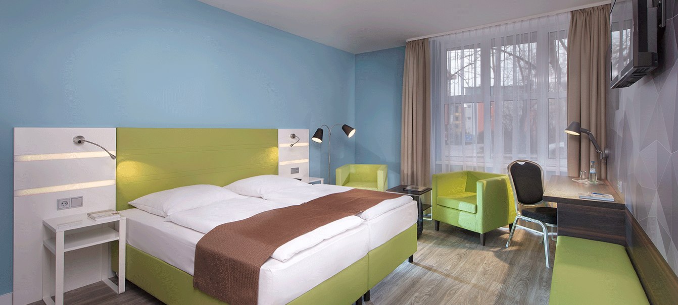 Modernes Doppelzimmer mit grünem Bett, Schreibtisch, zwei Sesseln und TV. Helle Wände und große Fenster sorgen für eine freundliche Atmosphäre., © TOMAS