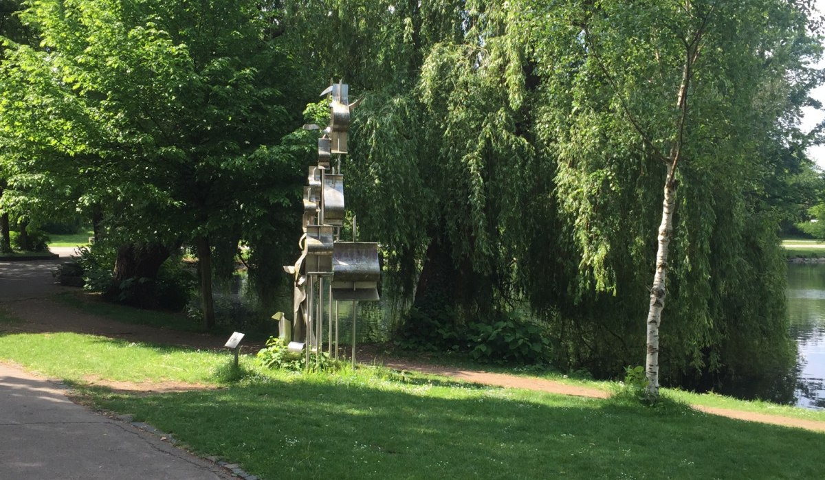 Eine Metallskulptur steht in einem grünen Park am Ufer eines Sees, umgeben von üppigen Bäumen und einem Gehweg., © www.pro-cycl.de Eine Metallskulptur steht in einem grünen Park am Ufer eines Sees, umgeben von üppigen Bäumen und einem Gehweg., © www.pro-cycl.de