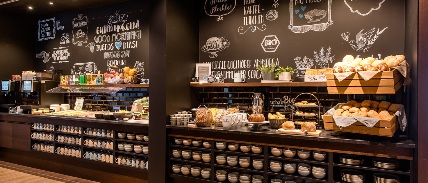 Ein Frühstücksbuffet mit Brotkörben, Tassen und Schüsseln vor einer dekorativen Kreidetafelwand., © TOMAS Ein Frühstücksbuffet mit Brotkörben, Tassen und Schüsseln vor einer dekorativen Kreidetafelwand., © TOMAS