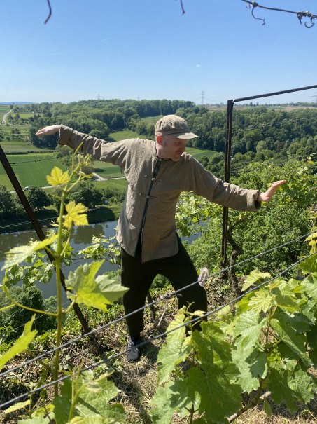 Person &uuml;bt Qigong in einem Weinberg &uuml;ber dem Neckar. Im Hintergrund sind gr&uuml;ne H&uuml;gel und ein Fluss zu sehen. Sonniger Tag., &copy; Karl Sch&uuml;le und Daniel Steinbauer