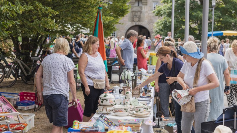Flohmarktbesucher st&ouml;bern an einem Stand mit Geschirr und Spielzeug. Im Hintergrund sind weitere Menschen und B&auml;ume zu sehen., &copy; BEBOP MEDIA