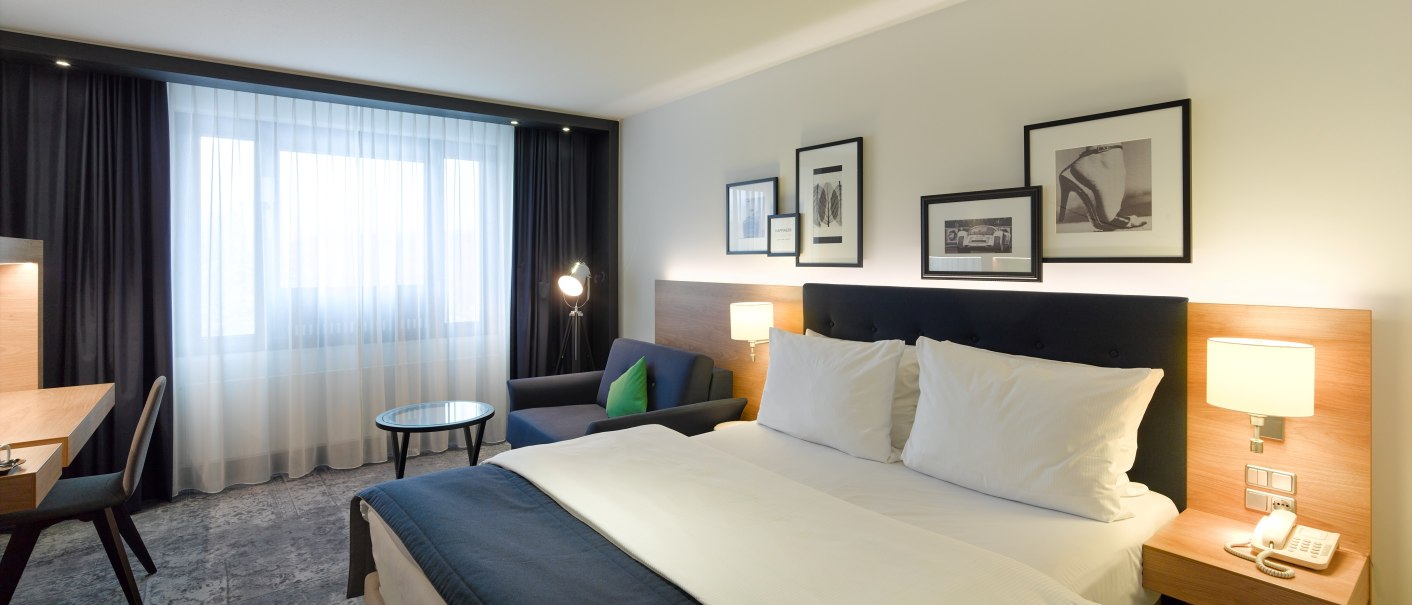 Modernes Hotelzimmer mit Doppelbett, Schreibtisch, Sofa und dekorativen Bildern an der Wand. Große Fenster mit Vorhängen lassen Tageslicht herein., © Holiday Inn Stuttgart Modernes Hotelzimmer mit Doppelbett, Schreibtisch, Sofa und dekorativen Bildern an der Wand. Große Fenster mit Vorhängen lassen Tageslicht herein., © Holiday Inn Stuttgart