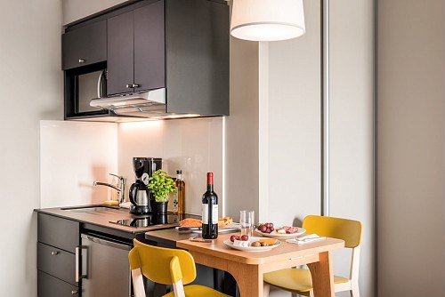 Kompakte Küche mit schwarzem Schrank, Esstisch, gelben Stühlen und Weinflasche auf dem Tisch., © adagio access Stuttgart Airport Messe