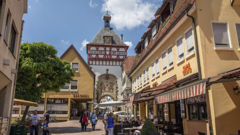 Eine belebte Altstadtstraße mit Fachwerkhaus und Uhrturm. Menschen sitzen in Straßencafés, während andere die Straße entlanggehen. Der Himmel ist blau., © Stuttgart Marketing GmbH Achim Mende