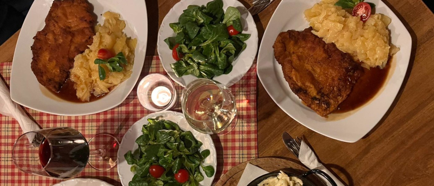 Zwei Teller mit Schnitzel, Kartoffelsalat und grünem Salat. Dazu Rot- und Weißwein auf kariertem Tischtuch., © Weinhof Zaiß
