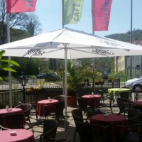 Café-Terrasse mit Tischen, roten Tischdecken und Sonnenschirmen. Im Hintergrund sind eine Straße, Gebäude und grüne Bäume zu sehen., © Intermezzo Café am Bach