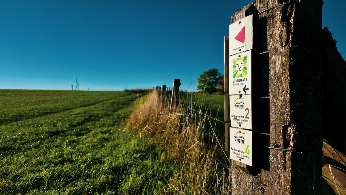Ein Holzpfosten mit Wanderschildern steht auf einer grünen Wiese. Der Himmel ist klar und blau, im Hintergrund sind Windräder sichtbar., © Landkreis Göppingen Ein Holzpfosten mit Wanderschildern steht auf einer grünen Wiese. Der Himmel ist klar und blau, im Hintergrund sind Windräder sichtbar., © Landkreis Göppingen