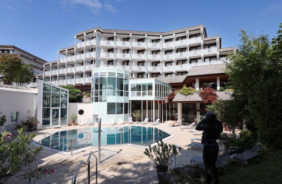 Ein modernes Hotel mit mehreren Etagen und Balkonen. Im Vordergrund ein Pool und eine Terrasse mit Liegest&uuml;hlen, umgeben von Pflanzen., &copy; Reha-Klinik Schw&auml;bische Alb