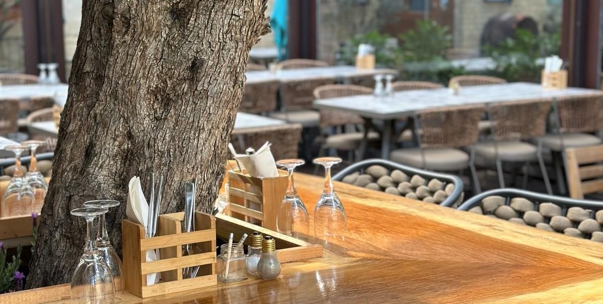 Ein Holztisch im Freien mit einem Baum in der Mitte, umgeben von Stühlen und gedeckten Tischen in einem Restaurant., © Meze Meze