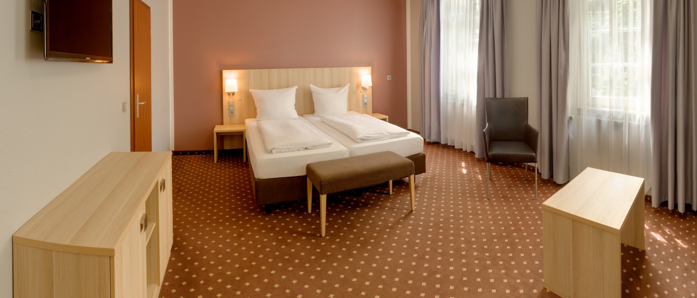 Geräumiges Hotelzimmer mit Doppelbett, braunem Teppich, Fernseher, Sessel und Tisch. Große Fenster mit Vorhängen lassen viel Licht herein., © attimo Hotel Stuttgart