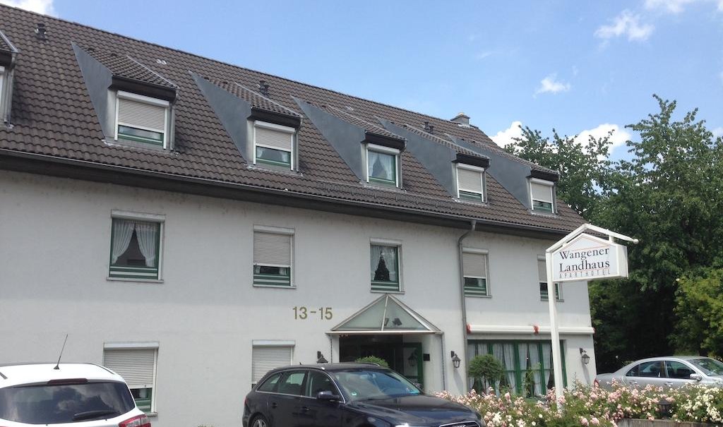 Das Aparthotel Wangener Landhaus mit grauem Dach und weißen Wänden. Vor dem Gebäude sind Autos geparkt. Ein Schild zeigt den Hotelnamen., © Aparthotel Wangener Landhaus