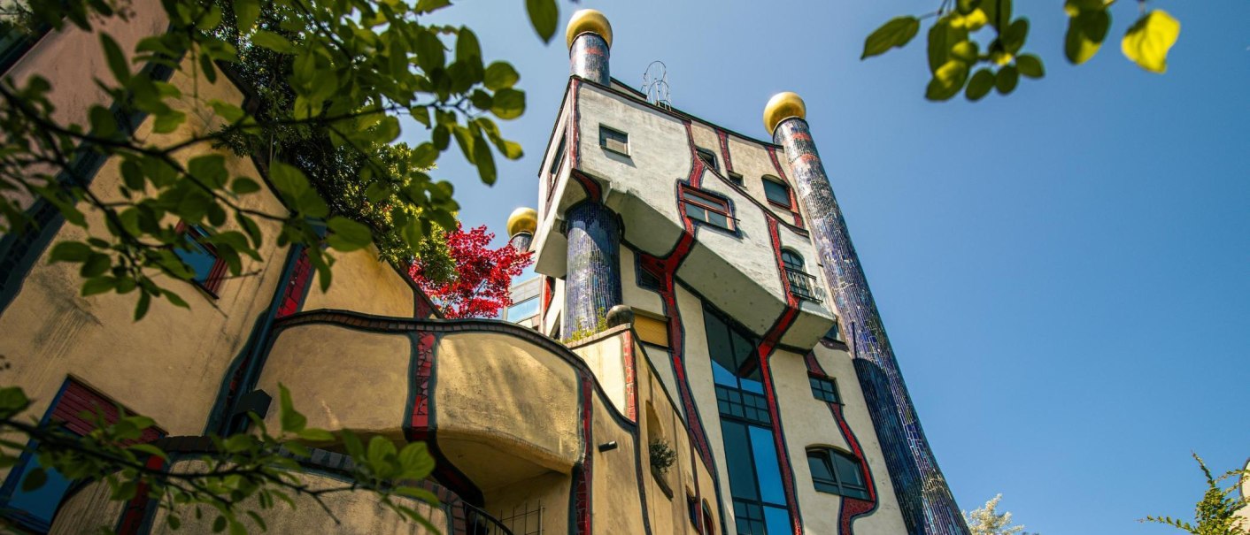 Das Hundertwasserhaus in Plochingen zeigt seine charakteristische bunte Fassade und organische Formen, umgeben von gr&uuml;nen Bl&auml;ttern und blauem Himmel., &copy; Stuttgart-Marketing GmbH, Sarah Schmid