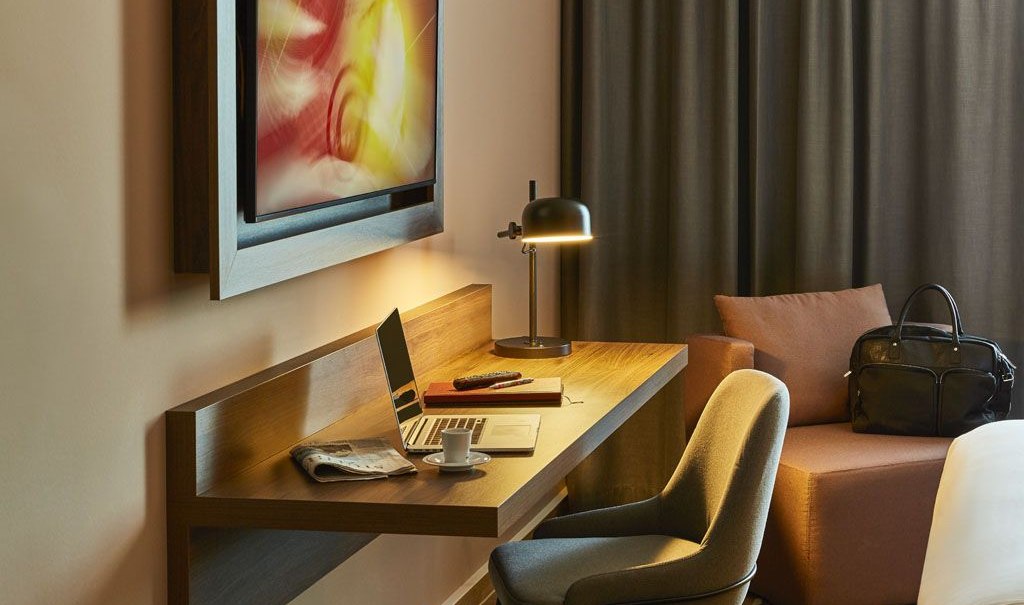 Modernes Hotelzimmer mit Schreibtisch, Laptop, Lampe und Sessel. Ein Kunstwerk hängt an der Wand, eine Tasche steht auf dem Sofa., © Pullman Stuttgart Fontana Modernes Hotelzimmer mit Schreibtisch, Laptop, Lampe und Sessel. Ein Kunstwerk hängt an der Wand, eine Tasche steht auf dem Sofa., © Pullman Stuttgart Fontana