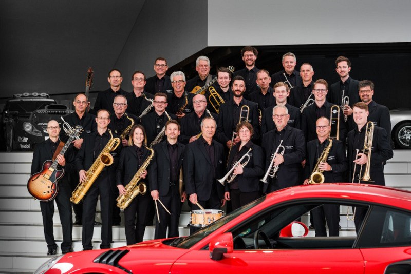 Eine Big Band mit verschiedenen Instrumenten posiert auf einer Treppe vor einem roten Porsche. Die Musiker tragen schwarze Kleidung., © Scala Kultur Live gGmbH Eine Big Band mit verschiedenen Instrumenten posiert auf einer Treppe vor einem roten Porsche. Die Musiker tragen schwarze Kleidung., © Scala Kultur Live gGmbH