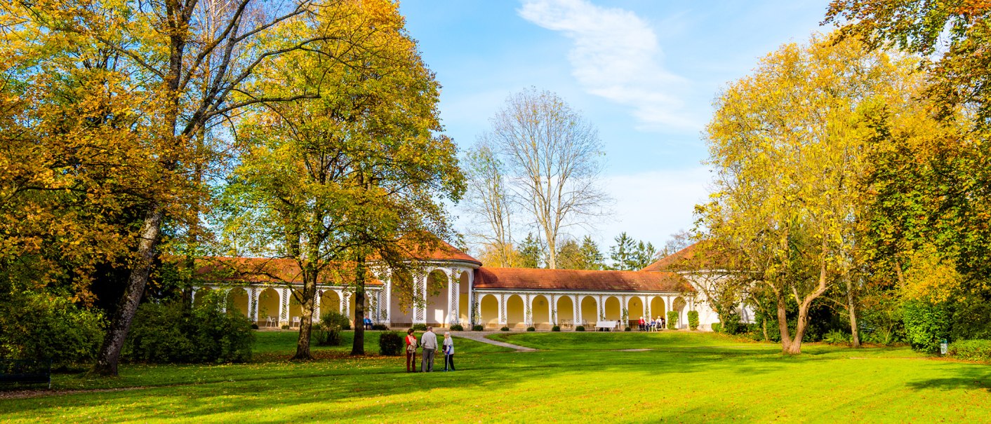 Herbstlicher Kurpark Bad Boll mit gelb-orange gefärbten Bäumen und einem historischen Gebäude mit Arkaden im Hintergrund., © Bad Boll Herbstlicher Kurpark Bad Boll mit gelb-orange gefärbten Bäumen und einem historischen Gebäude mit Arkaden im Hintergrund., © Bad Boll