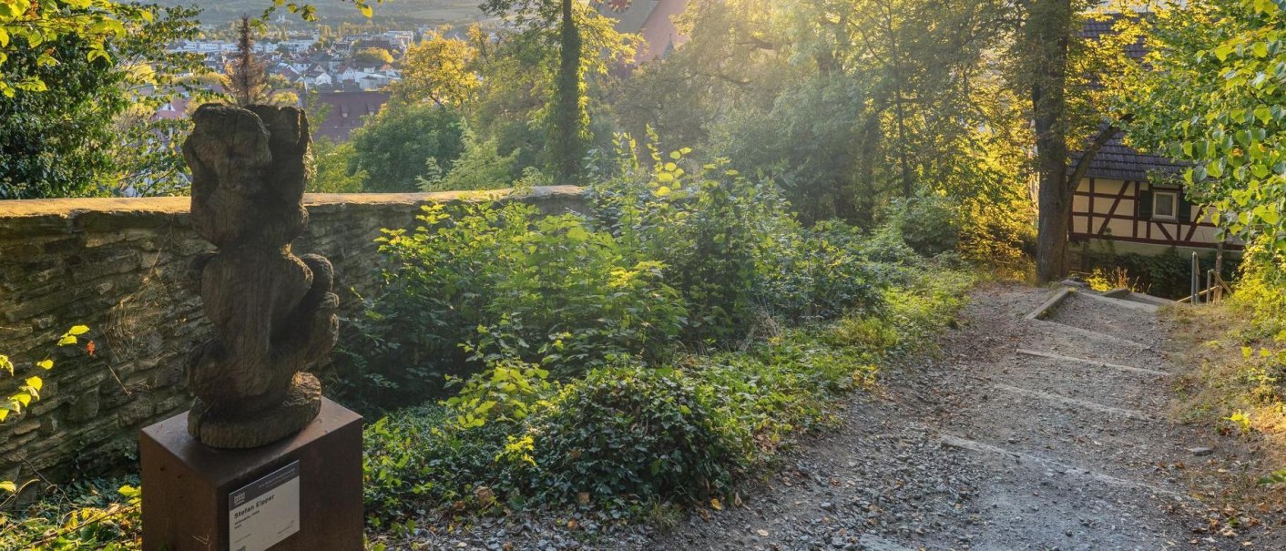 Skulpturenpfad in Herrenberg mit einer Holzskulptur, umgeben von B&auml;umen und einem Fachwerkhaus im Hintergrund. Die Abendsonne beleuchtet die Szene., &copy; Stuttgart-Marketing GmbH, Martina Denker