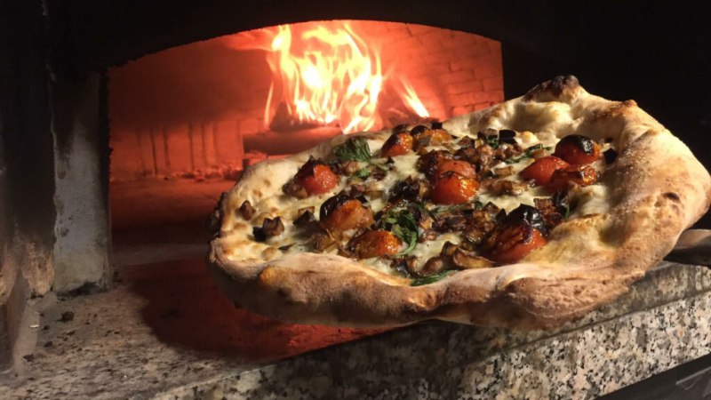 Eine frisch gebackene Pizza mit Tomaten und Kr&auml;utern wird aus einem hei&szlig;en Holzofen geholt. Der Ofen ist aus Stein und das Feuer lodert., &copy; DaCarmine