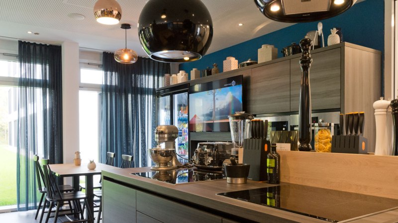 Moderne K&uuml;che mit Kochinsel, Esstisch, H&auml;ngelampen und Ger&auml;ten. Gro&szlig;e Fenster mit blauen Vorh&auml;ngen lassen viel Licht herein., &copy; Arthotel Ana / elaya