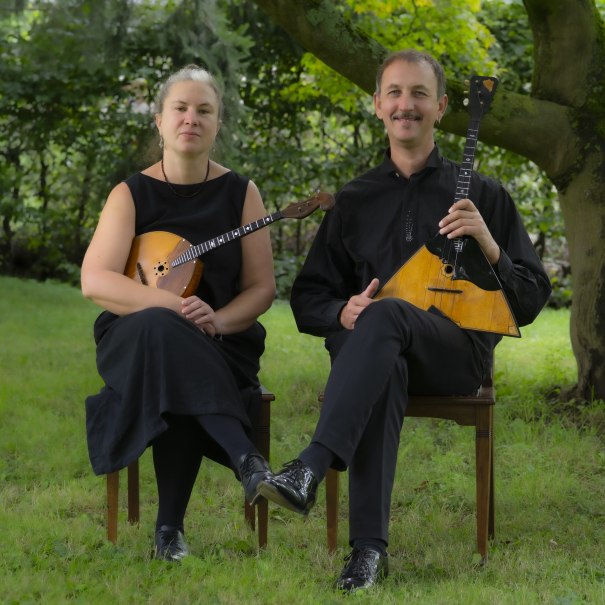 Zwei Musiker mit Balalaika und Domra sitzen im Freien auf St&uuml;hlen. Sie tragen schwarze Kleidung und sind von gr&uuml;ner Natur umgeben., &copy; Exprompt