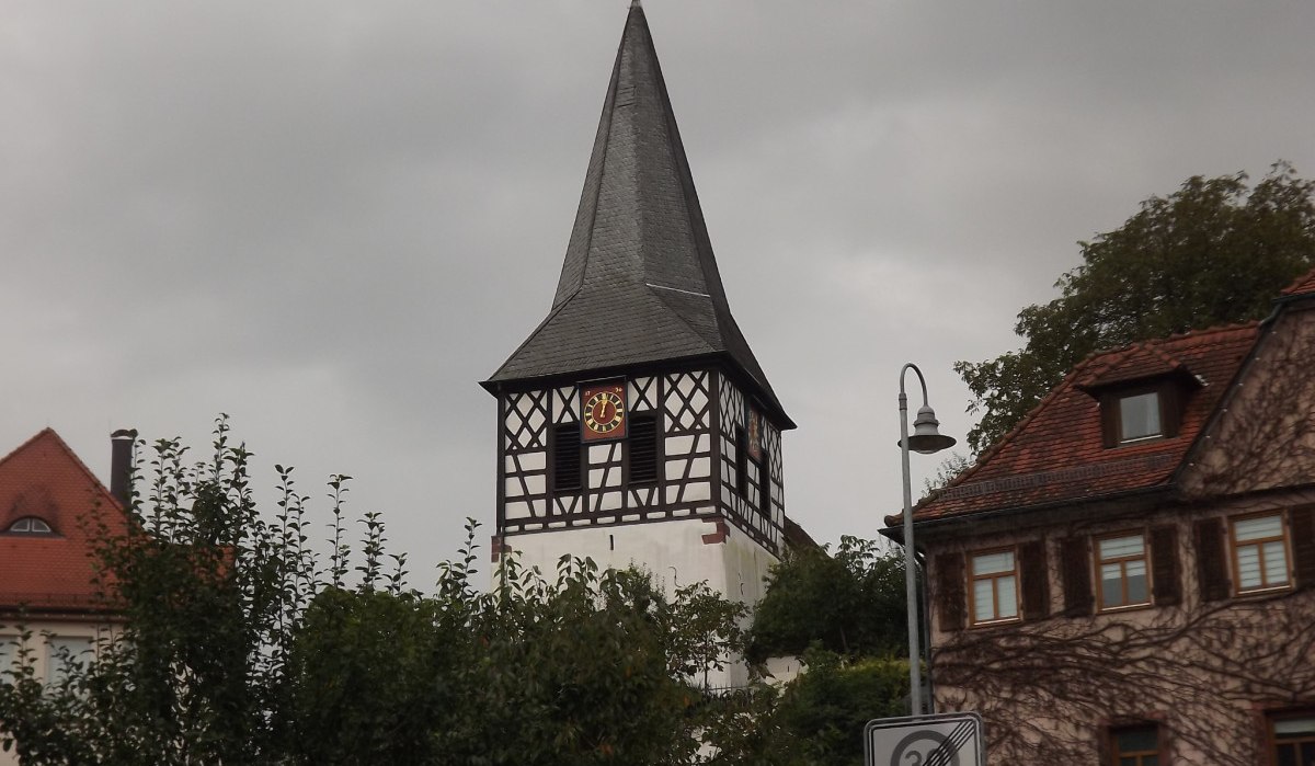 Ein Fachwerkturm mit Uhr und spitzem Dach erhebt sich vor einem bewölkten Himmel, umgeben von Bäumen und Häusern., © Natur.Nah. Schönbuch & Heckengäu Ein Fachwerkturm mit Uhr und spitzem Dach erhebt sich vor einem bewölkten Himmel, umgeben von Bäumen und Häusern., © Natur.Nah. Schönbuch & Heckengäu