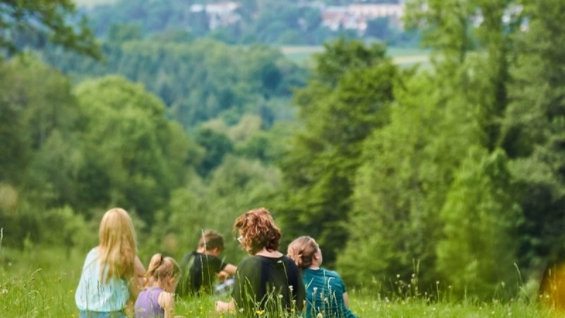 Gruppe von Menschen sitzt auf einer gr&uuml;nen Wiese, umgeben von B&auml;umen, mit Blick auf ein Dorf in der Ferne., &copy; Natur.Nah. Sch&ouml;nbuch & Heckeng&auml;u