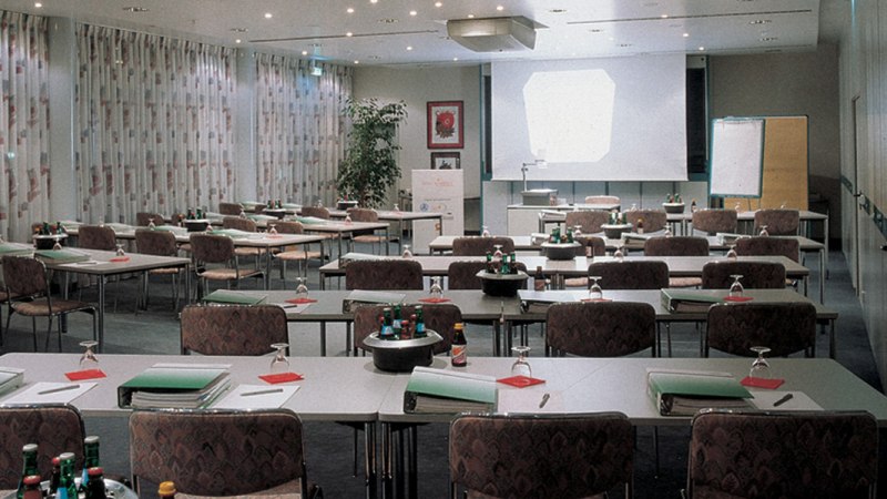 Ein leerer Konferenzraum mit Tischen, St&uuml;hlen, einem Projektor und einem Flipchart. Auf den Tischen liegen Ordner und Wasserflaschen., &copy; &copy; H-Hotels.com