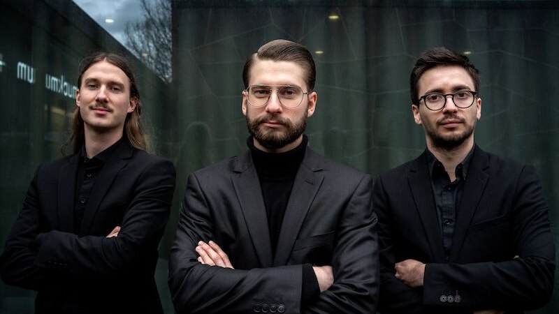 Drei Männer in schwarzen Anzügen posieren selbstbewusst vor einer modernen Glasfassade. Der Himmel ist bewölkt, was der Szene eine dramatische Note verleiht., © Interessengemeinschaft Kultur Sindelfingen / Böblingen e.V. Drei Männer in schwarzen Anzügen posieren selbstbewusst vor einer modernen Glasfassade. Der Himmel ist bewölkt, was der Szene eine dramatische Note verleiht., © Interessengemeinschaft Kultur Sindelfingen / Böblingen e.V.