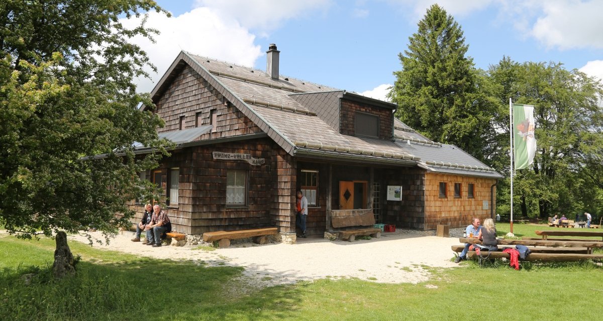 Das Franz-Keller-Haus ist ein rustikales Holzhaus mit Schindeldach, umgeben von Bäumen und Wiese. Menschen sitzen draußen und genießen die Umgebung., © Foto: Walter Laible Das Franz-Keller-Haus ist ein rustikales Holzhaus mit Schindeldach, umgeben von Bäumen und Wiese. Menschen sitzen draußen und genießen die Umgebung., © Foto: Walter Laible