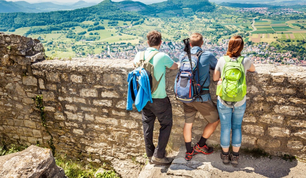 Drei Wanderer mit Rucksäcken stehen auf einer Mauer und genießen den Ausblick auf die grüne Landschaft von Hohenneuffen., © hochgehberge