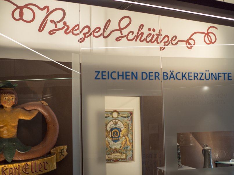 Ausstellung im Brezelmuseum mit dem Titel 'Brezelschätze'. Zu sehen sind Exponate der Bäckerzünfte, darunter ein Schild und ein Gemälde., © BrezelMuseum Ausstellung im Brezelmuseum mit dem Titel 'Brezelschätze'. Zu sehen sind Exponate der Bäckerzünfte, darunter ein Schild und ein Gemälde., © BrezelMuseum
