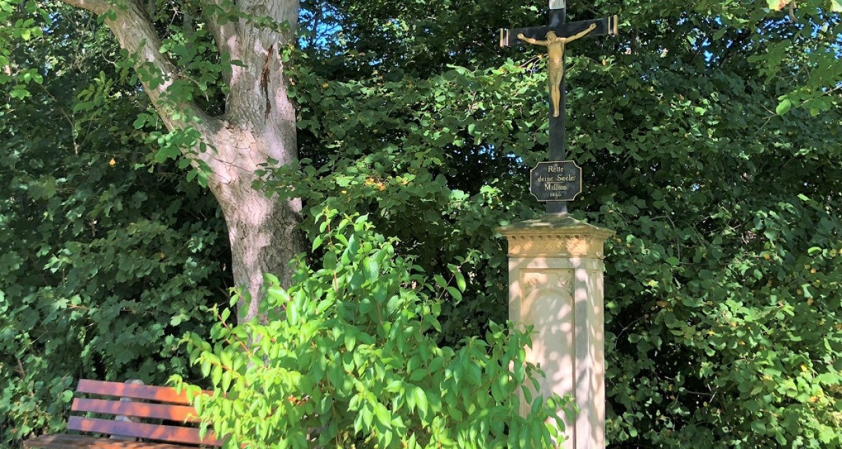 Ein steinernes Kreuz mit Jesusfigur steht neben einer Holzbank unter einem Baum, umgeben von dichtem Grün., © Touristik und Marketing GmbH Ein steinernes Kreuz mit Jesusfigur steht neben einer Holzbank unter einem Baum, umgeben von dichtem Grün., © Touristik und Marketing GmbH