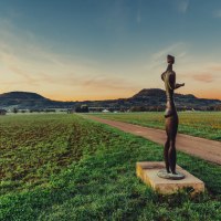 Eine Skulptur steht auf einem Feldweg, umgeben von grünen Feldern und Hügeln im Hintergrund, während die Sonne untergeht., © Landratsamt Ostalbkreis