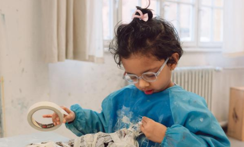 Ein Kind bastelt konzentriert mit Klebeband und Papier, tr&auml;gt eine blaue Sch&uuml;rze und eine Brille. Es sitzt an einem Tisch in einem hellen Raum., &copy; Kunstschule Labyrinth