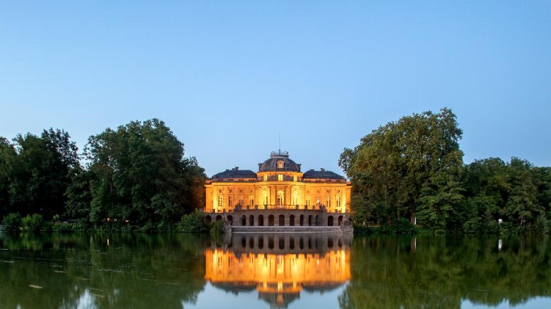 Das Seeschloss Monrepos erstrahlt in warmem Licht, umgeben von B&auml;umen und spiegelt sich im ruhigen Wasser wider. Der Himmel ist klar und blau., &copy; Seeschloss Monrepos &copy; Daniel Stauch