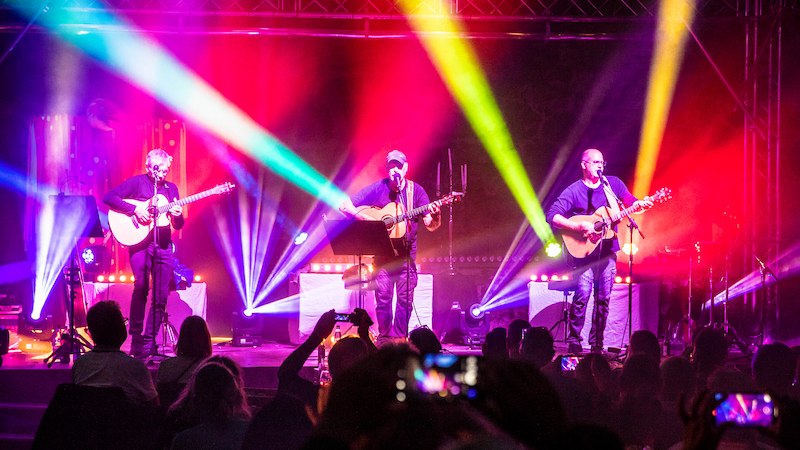 Drei Musiker mit Akustikgitarren auf einer B&uuml;hne, umgeben von bunten Lichteffekten. Das Publikum schaut zu und macht Fotos., &copy; Scala Kultur Live gGmbH