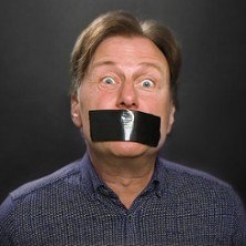 Ein Mann mit weit ge&ouml;ffneten Augen und schwarzem Klebeband &uuml;ber dem Mund vor einem dunklen Hintergrund., &copy; links im Bild