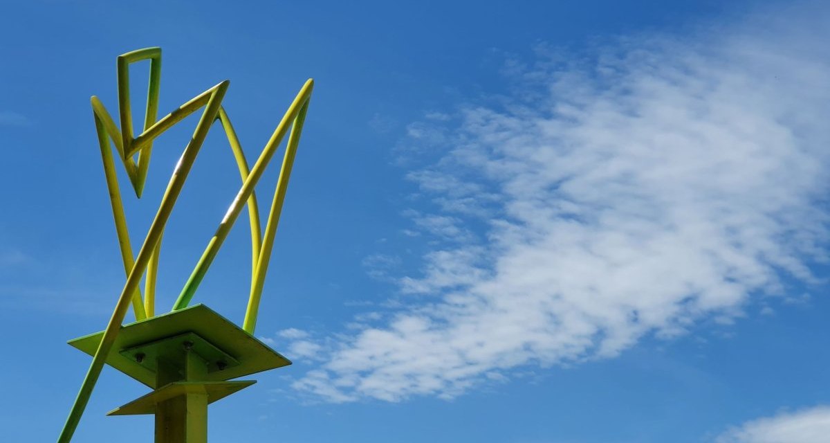 Eine gelbe, abstrakte Skulptur ragt in den blauen Himmel mit einigen Wolken. Im Hintergrund sind Bäume zu sehen. Eine gelbe, abstrakte Skulptur ragt in den blauen Himmel mit einigen Wolken. Im Hintergrund sind Bäume zu sehen.
