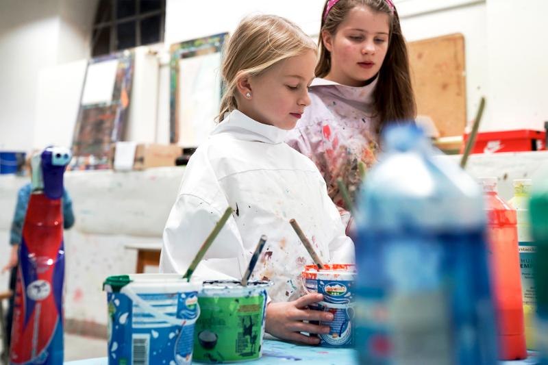 Zwei Kinder in einem Kunstworkshop, umgeben von bunten Farben und Pinseln. Sie tragen Malerkittel und sind konzentriert bei der Arbeit., © Kunstschule Labyrinth