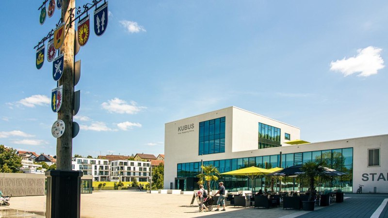 Moderner Marktplatz in Remseck mit dem Geb&auml;ude 'KUBUS', einem Maibaum mit Wappen und Menschen, die spazieren gehen. Sonniger Tag mit blauem Himmel., &copy; Stuttgart-Marketing GmbH, Sarah Schmid